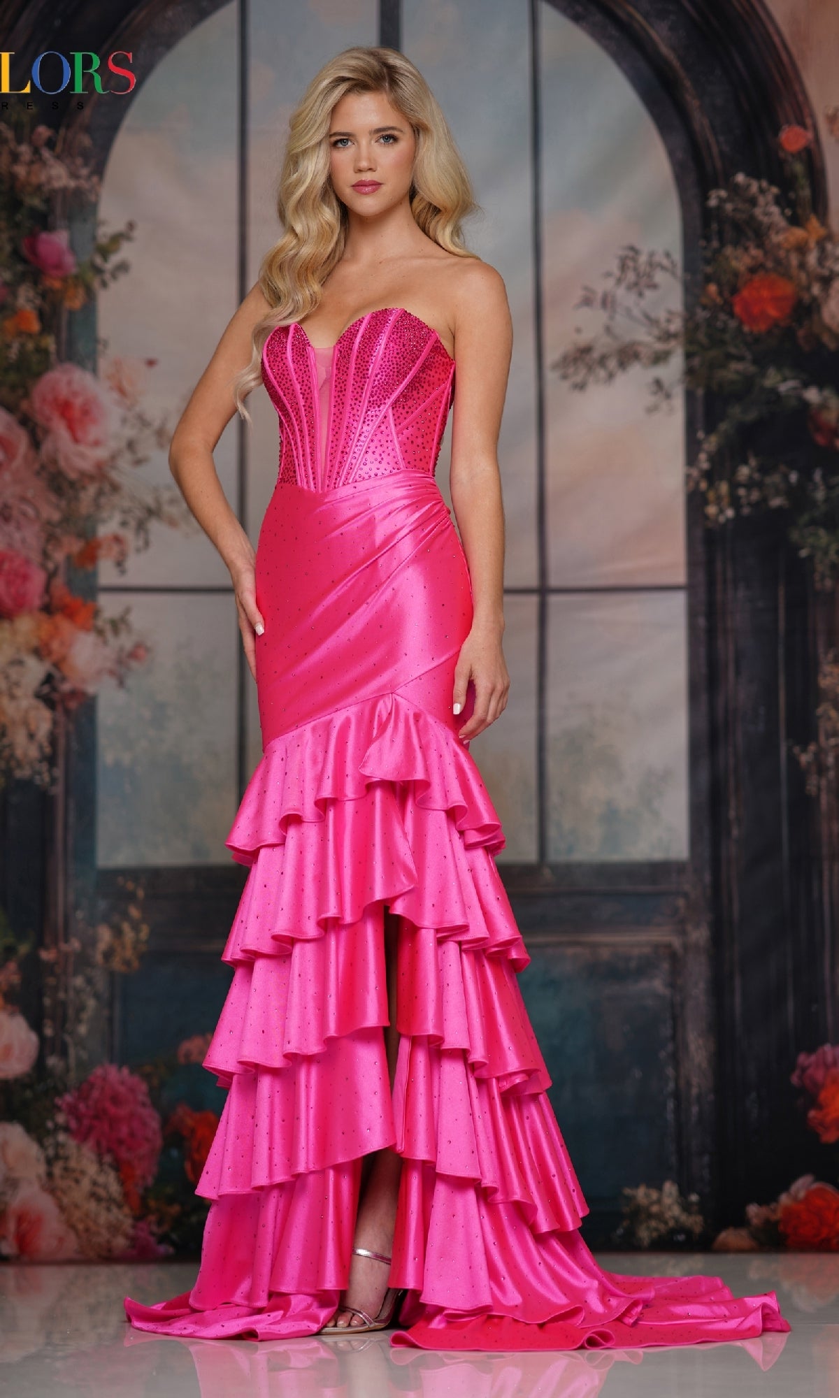 A woman stands indoors wearing the Colors Dress 3925, a long prom dress featuring a strapless bright pink corset-style fitted bodice and tiered skirt, set against a floral background with arched windows.